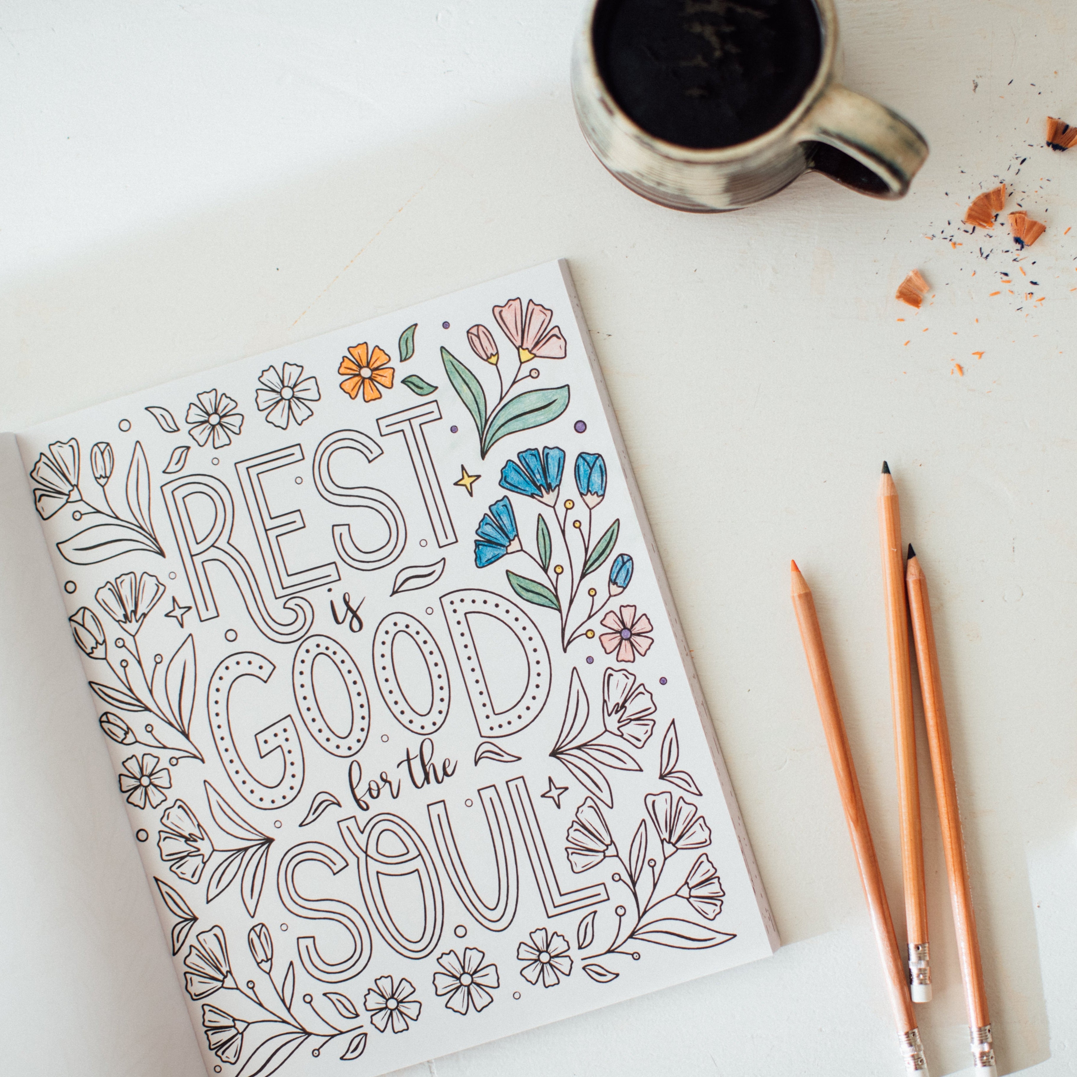 Coloring book with 'Rest is good for the soul' text and decorative elements on a white surface with a mug and pencils.